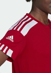 Czerwona sportowa koszulka z białymi paskami na ramionach, wykonana z gładkiego materiału. Posiada okrągły dekolt i logo Adidas na rękawie.