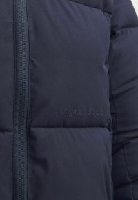 Marineblaue Steppjacke mit strukturierter Textur, ausgestattet mit einem Reißverschluss und geprägt mit dem "Originals Studio"-Logo auf dem Stoff.