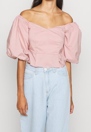Femme portant un haut rose clair à épaules dénudées avec des manches bouffantes, associé à un jean bleu clair taille haute, visible des épaules aux hanches.