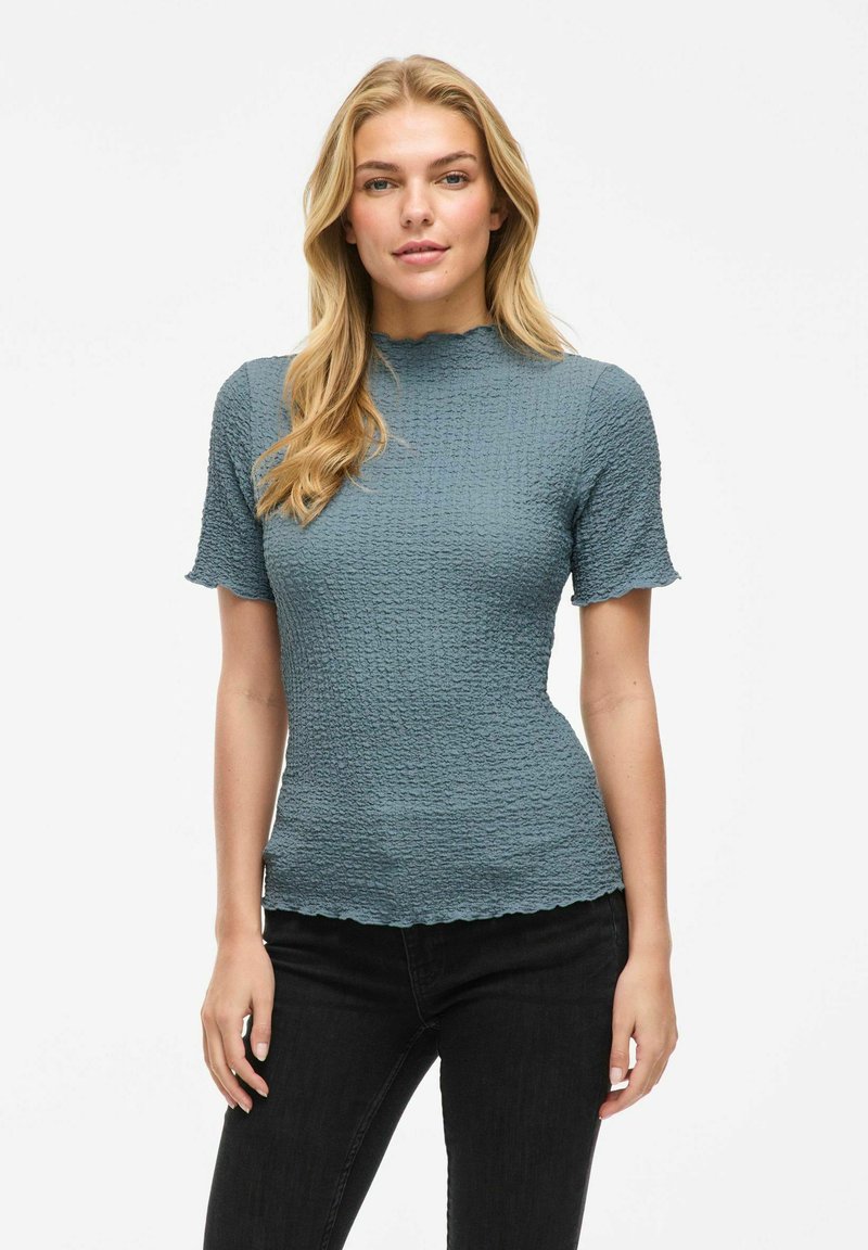 Top corto a maniche corte in blu opaco, con collo alto e orlo ondulato. Indossato con jeans neri.