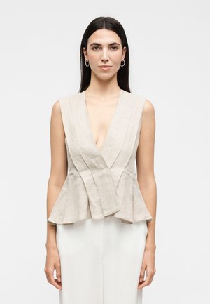 Kvinde med lige mørkt hår iført ærmeløs beige slå-om-top med plisseringer og hvide bukser, stående mod en ensfarvet lys baggrund.