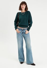Pull court vert avec des poignets côtelés, associé à un jean flare bleu clair. La modèle porte également des bottes à talons noires et un sac à épaule.