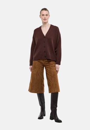 Femme vêtue d’un cardigan marron foncé boutonné avec col en V, d’un pantalon large court en daim marron et de bottes noires hautes à talons, debout face à l’avant.
