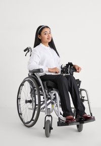 Rollstuhl mit einem silbernen Metallrahmen, schwarzem Sitz, Benutzer in einem weißen Hemd, schwarz gestreiften Hosen und kastanienbraunen Schuhen, der eine schwarze Tasche hält.