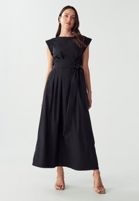 Vestito nero senza maniche con corpetto aderente, gonna plissettata e cintura annodata in vita. Tessuto liscio e design lungo fino al pavimento. Sfondo neutro.