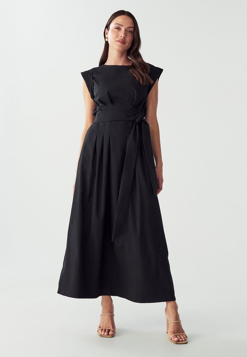 Vestito nero senza maniche con corpetto aderente, gonna plissettata e cintura annodata in vita. Tessuto liscio e design lungo fino al pavimento. Sfondo neutro.