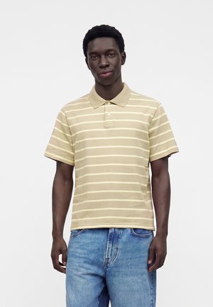 Mann, der ein beige-weiß gestreiftes kurzärmeliges Poloshirt und blaue Jeans trägt, steht vor einem einfarbig weißen Hintergrund.
