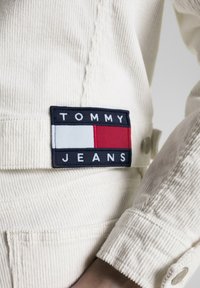 Chaqueta de pana blanca con un parche azul marino oscuro. El parche presenta "TOMMY JEANS" en letras blancas y rectángulos de colores en rojo y azul claro.