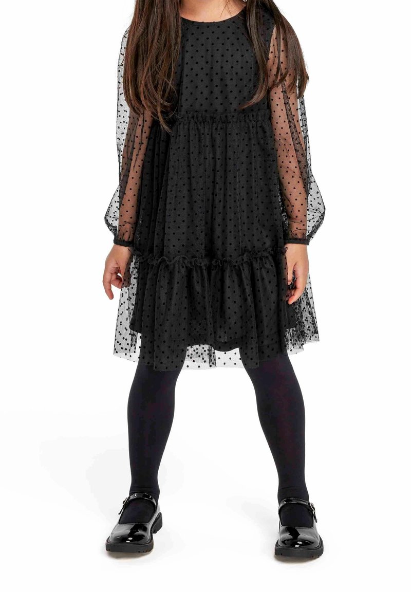 Jeune fille portant une robe noire transparente à pois avec manches longues, collants noirs et chaussures Mary Jane noires brillantes, debout, les jambes écartées.