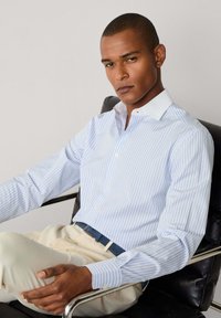 Uomo che indossa una camicia a righe bianca e azzurro chiaro e pantaloni beige, seduto su una sedia in pelle nera contro uno sfondo semplice.