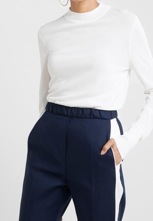 Personne portant un haut ajusté à manches longues blanc et un pantalon taille haute bleu marine avec des bandes blanches sur les côtés, la main dans la poche.