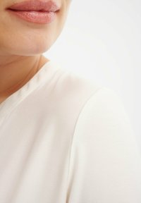Close-up van een gladde, lichtbeige stof met een subtiele glans bij de halslijn, die een zacht, schoon ontwerp en minimale stikdetails laat zien.