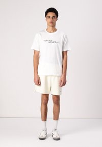 Camiseta de algodón blanca con "JACK & JONES" impreso en gris, combinada con pantalones cortos color crema claro. Modelo lleva calcetines blancos y zapatillas grises.