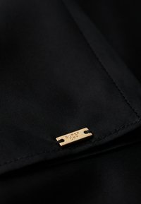 Tissu noir au toucher lisse, arborant une petite étiquette en métal doré gravée avec "SUPER DRY" et attachée par deux trous.