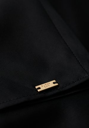 Tejido negro con una textura suave, que presenta una pequeña etiqueta de metal dorado grabada con "SUPER DRY" y sujeta por dos agujeros.