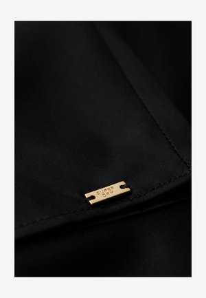 Tejido negro con una textura suave, que presenta una pequeña etiqueta de metal dorado grabada con "SUPER DRY" y sujeta por dos agujeros.