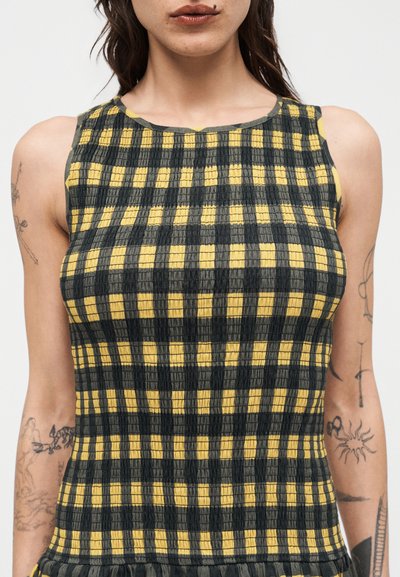 Γυναίκα που φορά αμάνικο φανελάκι με ριγέ υφή και κίτρινο και σκούρο πράσινο καρό μοτίβο, με εμφανή τατουάζ και στα δύο χέρια.