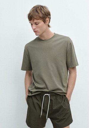 Giovane uomo con capelli castano chiaro corti, che indossa una maglietta verde oliva e pantaloncini verde scuro con coulisse bianca, in piedi con le mani in tasca.