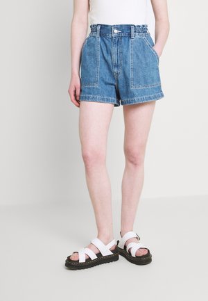 Persoon draagt een high-waisted blauwe spijkershorts met grote voorzakken en witte sandalen met zwarte zolen.