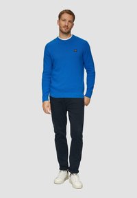 Blauwe gebreide trui met een ribbelpatroon, ronde halslijn en een klein logo-detail. Gecombineerd met donkere broek en witte sneakers.