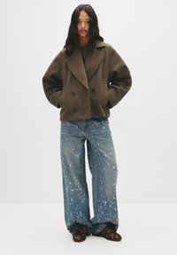 Manteau en laine marron à double boutonnière avec de grands revers, associé à un jean léger bleu clair à jambes larges présentant des motifs éclaboussés et des chaussures noires à motif animal.