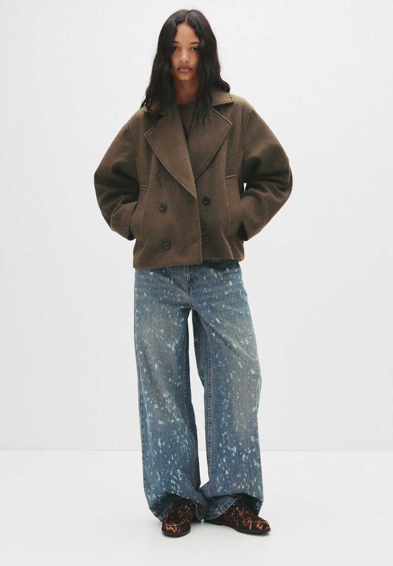 Manteau en laine marron à double boutonnière avec de grands revers, associé à un jean léger bleu clair à jambes larges présentant des motifs éclaboussés et des chaussures noires à motif animal.