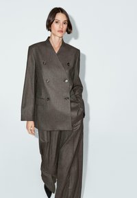 Abito marrone doppiopetto realizzato in tessuto testurizzato, con quattro bottoni e due tasche laterali, abbinato a pantaloni larghi e scarpe nere.