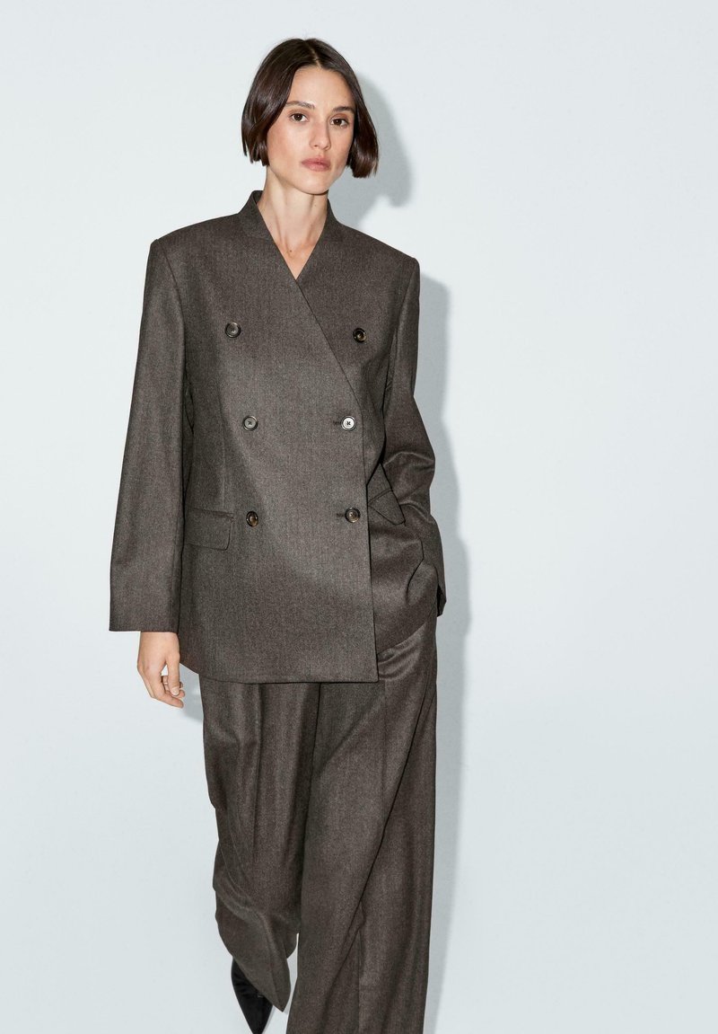 Abito marrone doppiopetto realizzato in tessuto testurizzato, con quattro bottoni e due tasche laterali, abbinato a pantaloni larghi e scarpe nere.