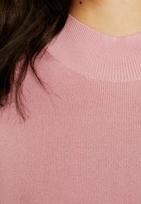 Pull en tricot rose clair avec un col rond côtelé. La texture est lisse, présentant un motif uniforme sur tout le tissu.