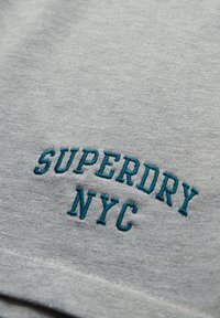 Sudadera de algodón gris con el texto bordado en azul "SUPERDRY NYC" en una fuente negrita y ligeramente arqueada en el centro. Textura suave.