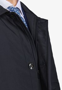 Sort jakke med en glat overflade, featuring en lynlåslukning og knapdetalje. Underneath ses en lyseblå skjorte med et mønstret slips.