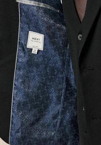 Schwarzes Sakko mit einem gepunkteten marineblauen Futter, das geometrische Formen zeigt. Das Etikett zeigt "NEXT Tailoring, Slim Fit, 40R, Hergestellt in Vietnam."