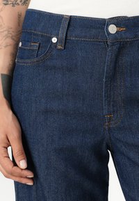 Mørkeblå denimjeans med oransje søm, med knappelukking foran, beltehemper og sidelommer. Jeansen har en glatt tekstur.