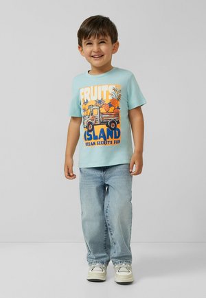Smiling young boy standing, wearing light blue t-shirt with colorful truck and fruit design, light jeans, and white sneakers.