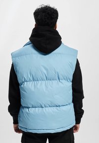 Lysblå puffervest med høj krave og ærmeløs design, med quiltede sektioner og en glat, shiny tekstur. Sort hættetrøje under.