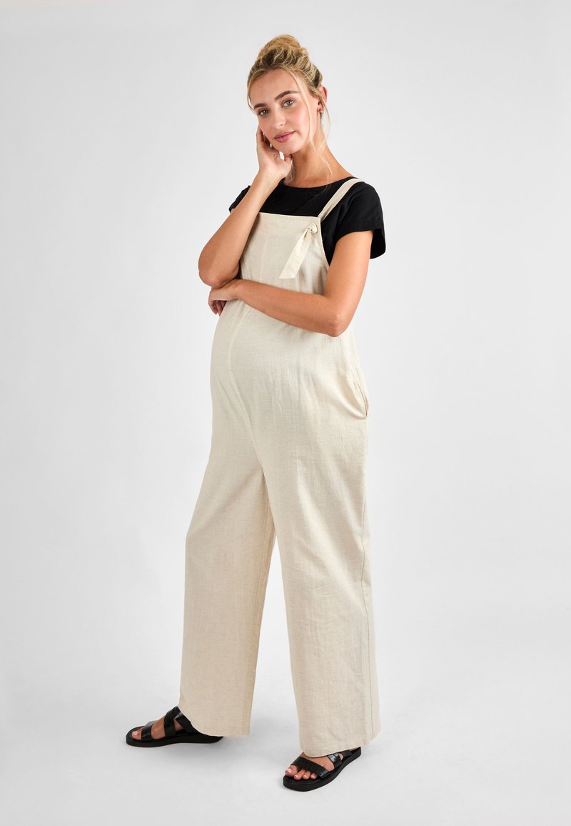 Gravid kvinde med blond hår sat i en knold bærer beige overalls og sorte sandaler, poserer med den ene hånd på ansigtet og den anden på maven.