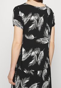 Černá blůza s krátkými rukávy, která má bílý listový vzor, kulatý výstřih a hladkou texturu látky. Zobrazeno zezadu.