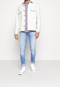 Hellgraue Knopfjacke mit zwei Brusttaschen über einem lila karierten Hemd, kombiniert mit hellblauen Jeans und weißen Sneakern mit hohem Schnitt.