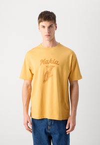 Gul bomull t-skjorte med brun grafisk trykk av en fugl og "Makia" over. Korte ermer, rund hals, løs passform.