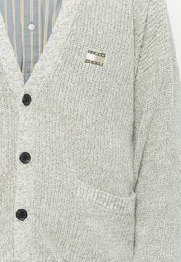 Gray knitted cardigan featuring a V-neck, black buttons, diagonal ribbed texture, and a rectangular "Tommy Jeans" logo patch on the chest.
