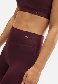 Sujetador deportivo burdeos y leggings de tiro alto hechos de un material suave y elástico. Presenta costuras sutiles y un logotipo en la cintura.