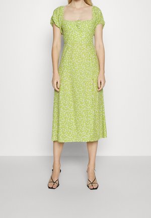 Kobieta stojąca w zielonej sukience midi w białe kwiaty, z bufiastymi rękawami, kwadratowym dekoltem i czarnymi sandałkami na wysokim obcasie.