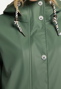 Veste imperméable verte avec cordons rayés et perles en bois noir, ornée de boutons-pression blancs à logo sur le devant.
