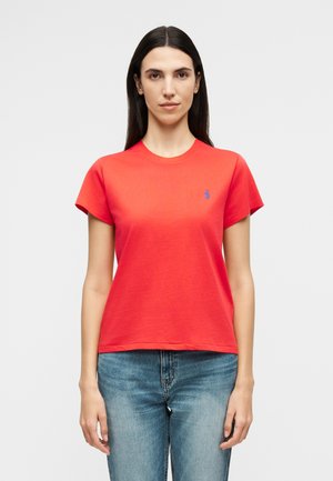 Pikkade tume juustega naine, kes kannab eredat punast lühikeste varrukatega T-särki ja siniseid teksapükse, tavalise heleda tausta ees.