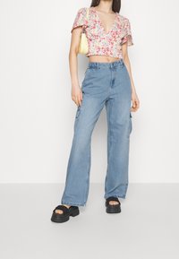 Top corto floral con escote en V profundo y mangas con volantes. Pantalones cargo de talle alto en un azul claro con bolsillos laterales. Sandalias de plataforma negras.