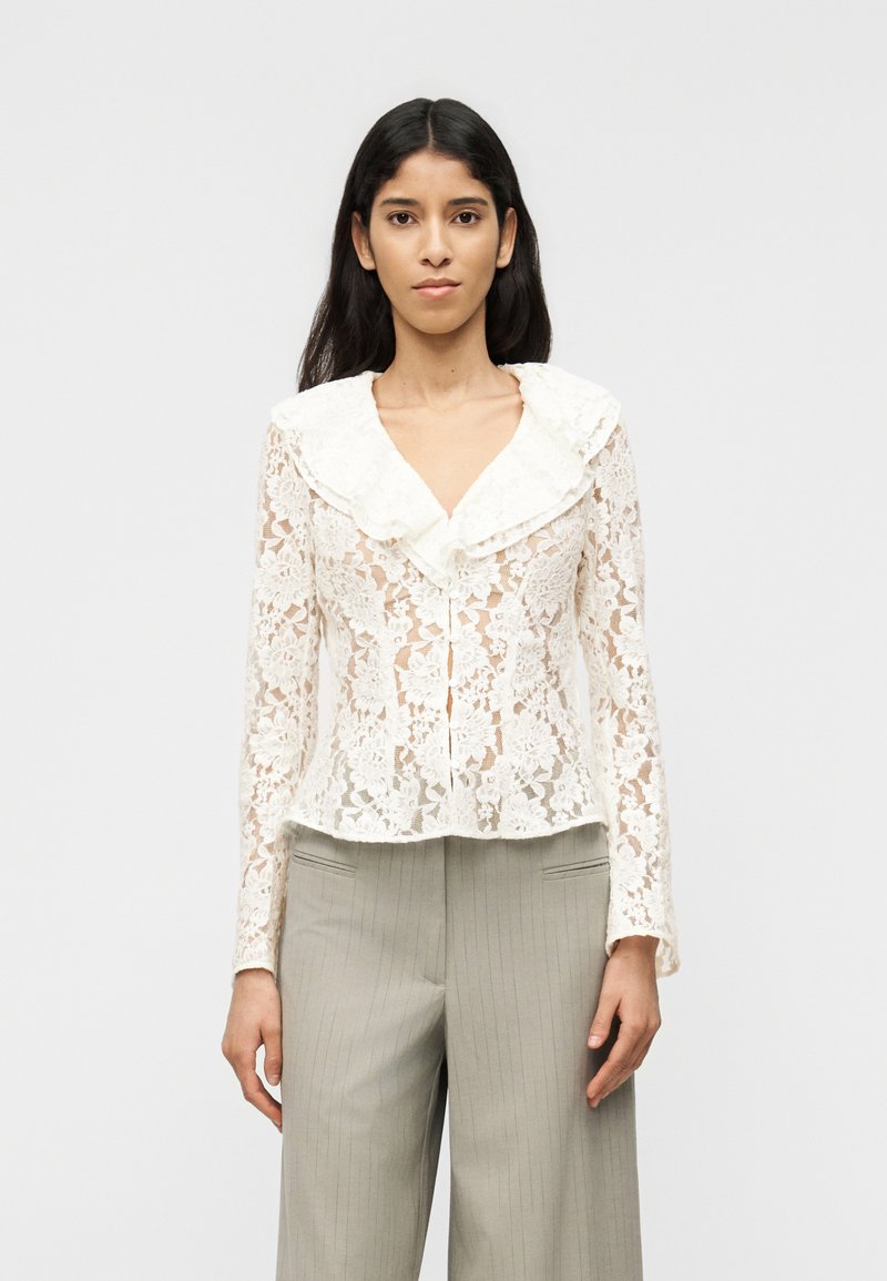 Γυναίκα που φοράει διαφανή λευκή δ αντελένια μπλούζα με μεγάλο βολάν στον γιακά και φαρδιά γκρι παντελόνια με λεπτές ρίγες.