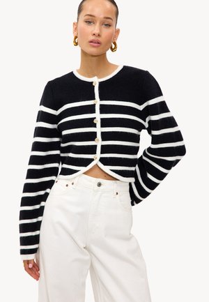 Cardigan rayé noir et blanc avec une silhouette courte, doté d'un col rond et de boutons dorés proéminents, associé à un pantalon blanc.