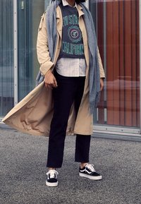 Gabardina beige sobre un sudadera gris con texto verde, combinada con jeans negros y zapatillas negras con suelas blancas. Bufanda gris clara.