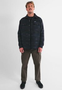 Hombre de pie mirando al frente, vestido con una camisa de botones negra con estampado, pantalones cargo marrones y zapatillas negras, contra un fondo blanco liso.