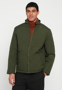 Casaco verde acolchoado com gola alta, apresentando um fecho de correr frontal e bolsos laterais. Usado sobre uma camisola de gola alta castanha. Textura lisa.